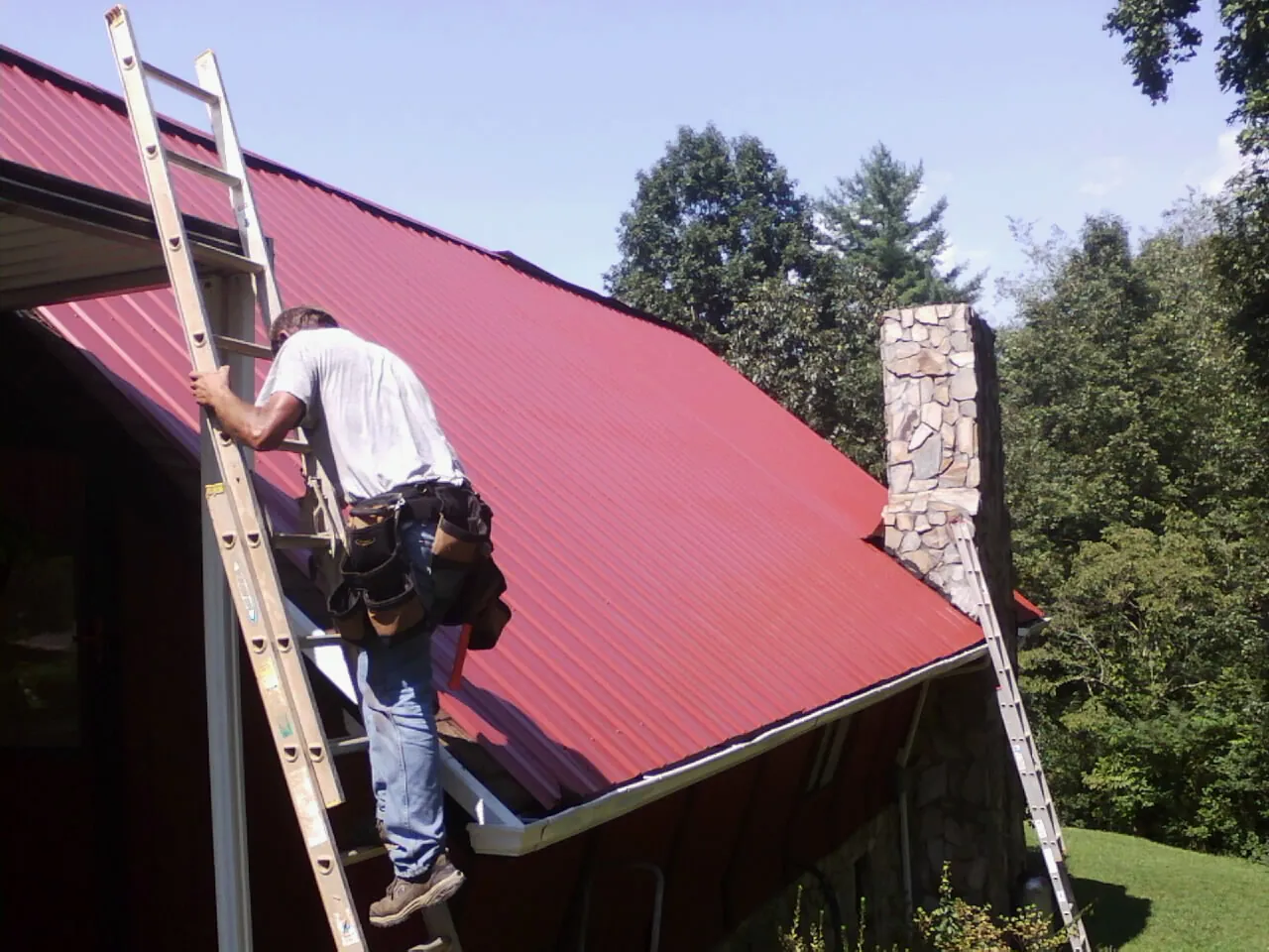 Roofer on a ladder beside a red metal roof and stone chimney — Soffit & Fascia Repair by Roofing Brothers in Ellijay